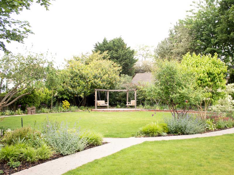 large suburban garden with central curved lawn and lush planting throughout. A sturdy swing set is positioned near the hedge line