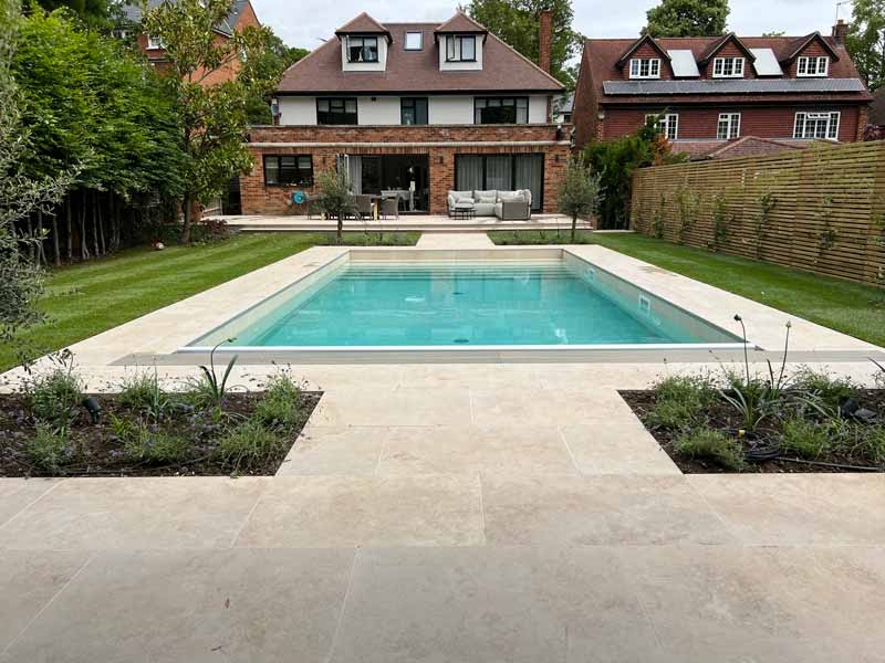 English garden with rectangular swimming pool surrounded by jura beige porcelain paving which matches adjoining patio