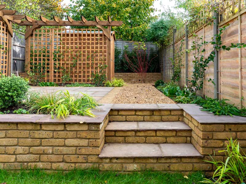 a low brick retaining wall is pierced by a set of garden steps which lead to a trellis-framed terrace