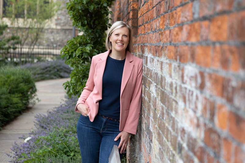 garden designer julia langsman wearing blue jeans with a pink jacket and leaning on a brick wall