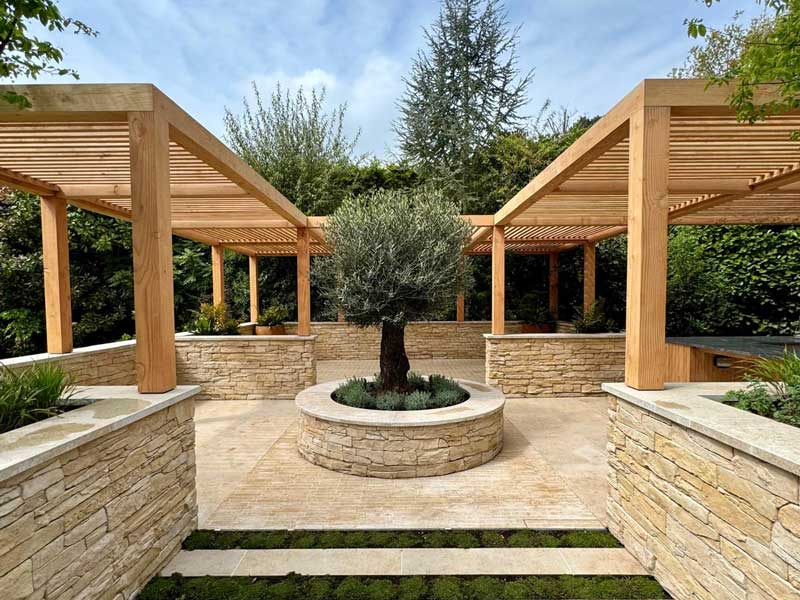 beautiful mediterranean style paved garden with timber pergola and central olive tree planted in a raised bed with bespoke stone coping stones